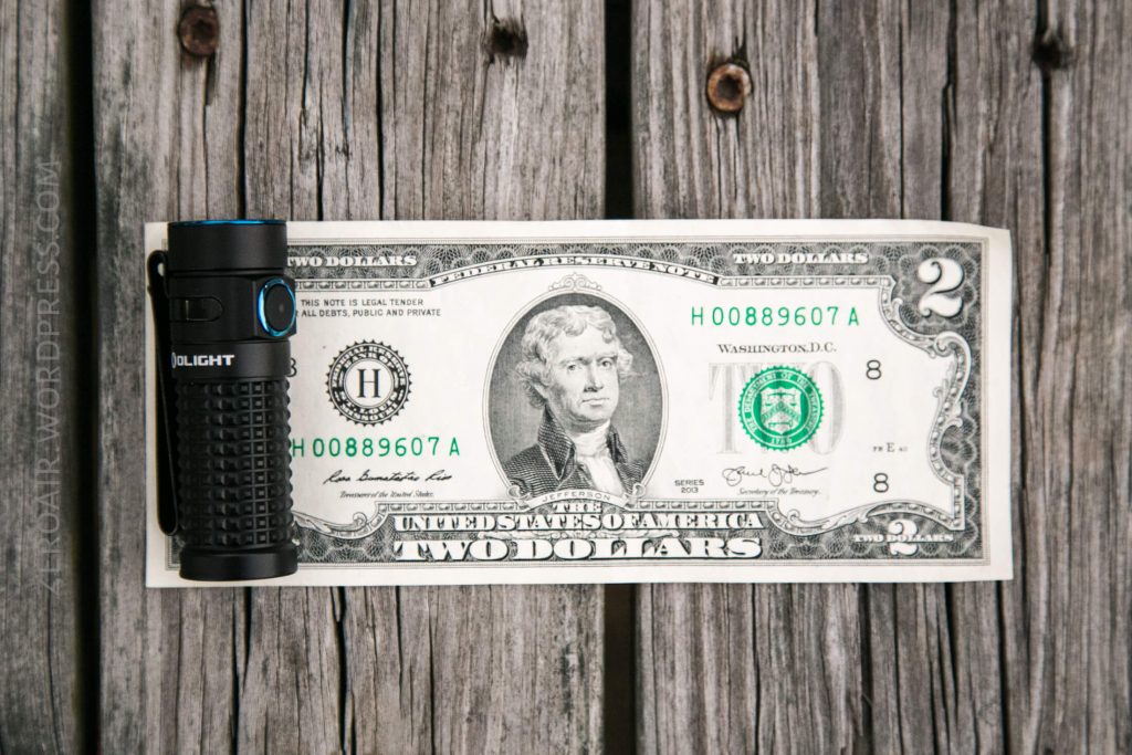 A black Olight flashlight is placed vertically on the left side of a two-dollar bill, which is laid flat on a weathered wooden surface.