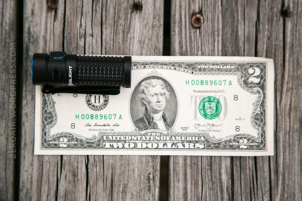 A black flashlight is placed on top of a two-dollar bill, both lying on a weathered wooden surface. The bill features a portrait of Thomas Jefferson.