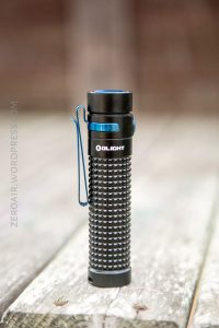 A black and blue Olight flashlight with a textured grip and clip is standing upright on a wooden surface outdoors. The background is blurred.