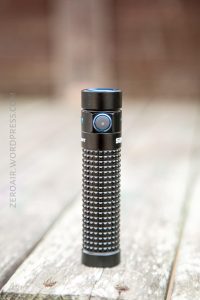 A black, textured flashlight standing upright on a weathered wooden surface with a blurred brown background. The flashlight has a round button near its top.