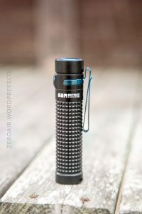 A compact black flashlight with textured grip, labeled S2R BATON II, stands upright on a weathered wooden surface. A blue-edged pocket clip is attached to the side.