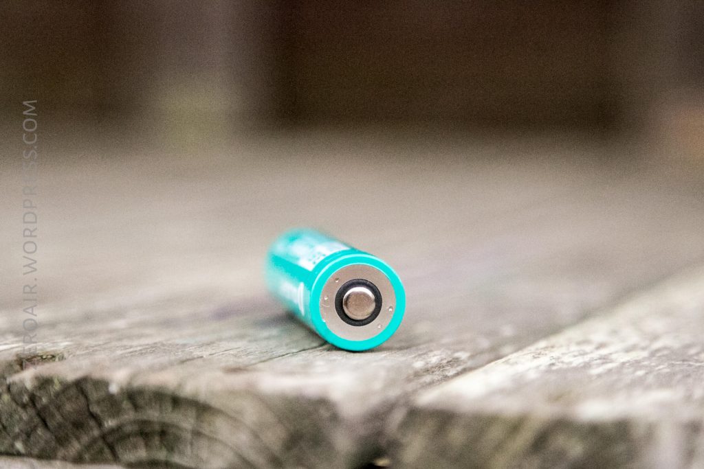 A turquoise AA battery lies horizontally on a weathered wooden surface, with the battery’s positive end facing the camera. The background appears blurred.