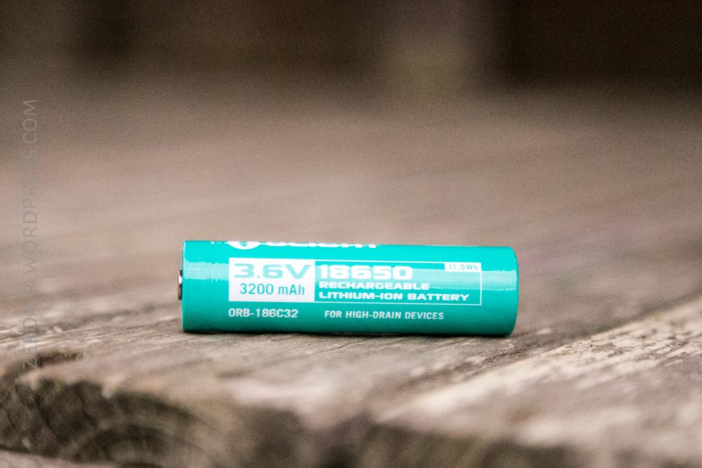 A turquoise Olight 18650 rechargeable lithium-ion battery with 3.6V and 3200mAh capacity lies on a wooden surface. The battery text includes for high-drain devices and product information.