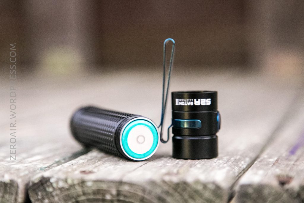 A small black flashlight is shown on a wooden surface with its battery compartment open and the tailcap detached, revealing the battery inside. The flashlight has a textured grip and a blue ring around the battery contact.