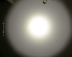 A bright circular light is projected onto a wall, creating a central white spot with a gradual dark halo. The text ZEROAIR.ORG is visible vertically on the left side. Part of a ceiling fixture appears at the top.