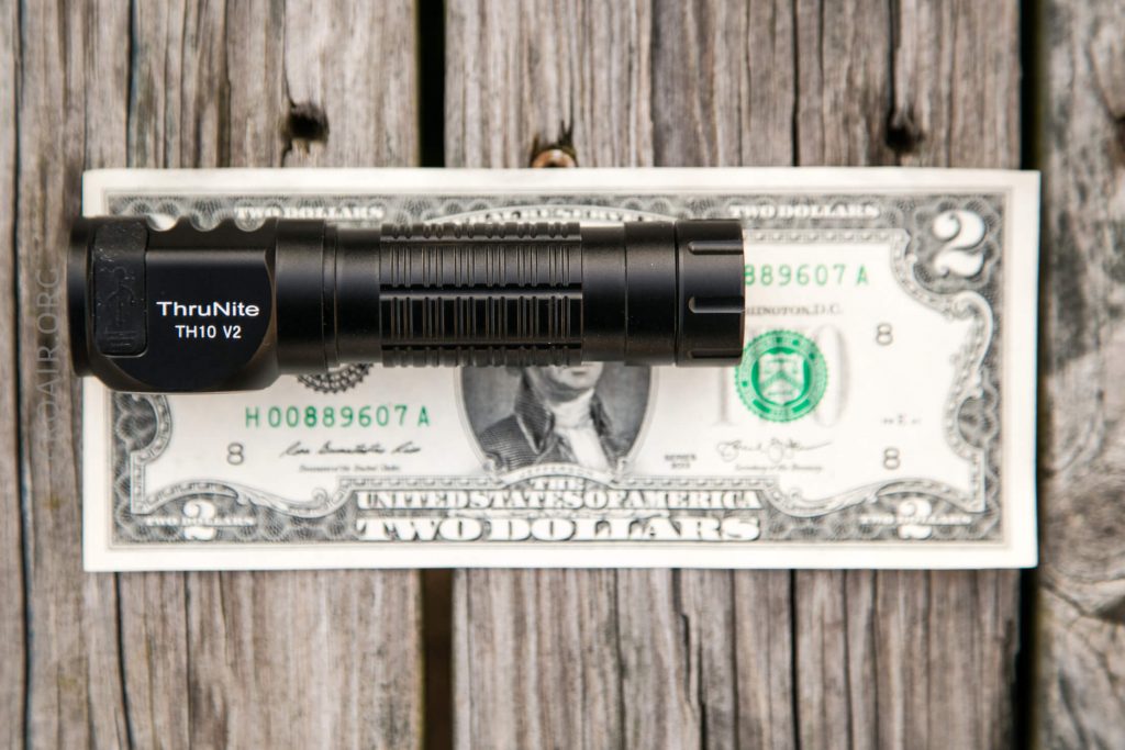 A black ThruNite TH10 V2 flashlight is placed horizontally on top of a two-dollar bill resting on a weathered wooden surface.