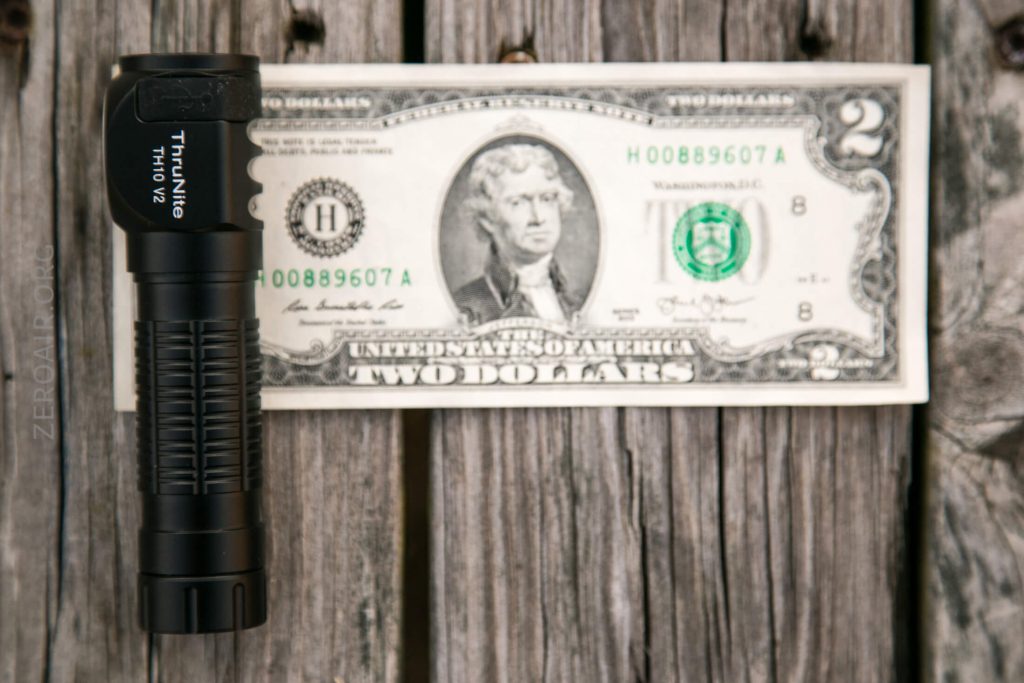 A black ThruNite TNV12 flashlight is placed vertically next to a two-dollar bill lying flat on a weathered wooden surface. The bill displays Thomas Jefferson’s portrait and the number 2.