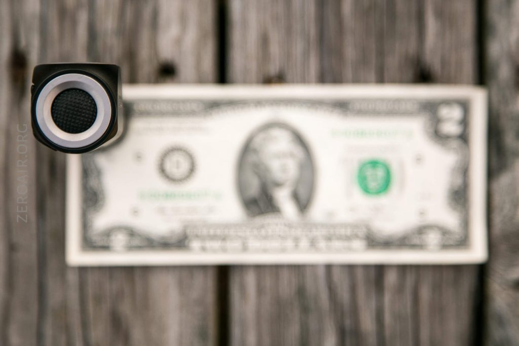 A close-up of a gun barrel pointed toward the camera with a blurred two-dollar bill laying flat on a wooden surface in the background.
