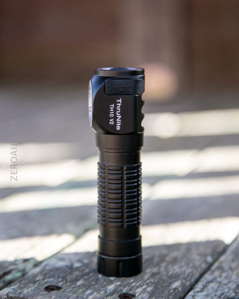 A black ThruNite TH10 V2 flashlight stands upright on a weathered wooden surface with blurred sunlight and shadows in the background.