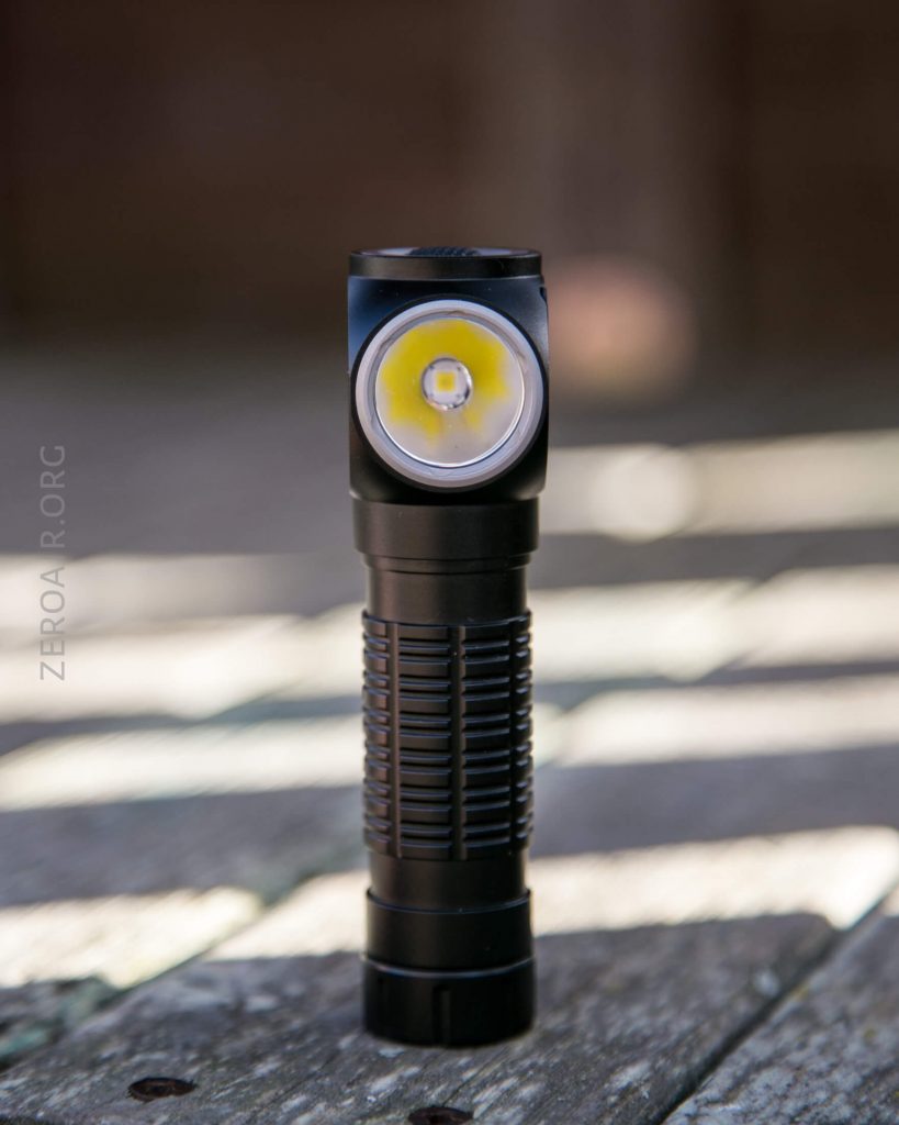 A black flashlight stands vertically on a wooden surface outdoors, with a bright yellow LED visible in the lens. The words ZEROAIR.ORG are faintly visible on the left side of the image.