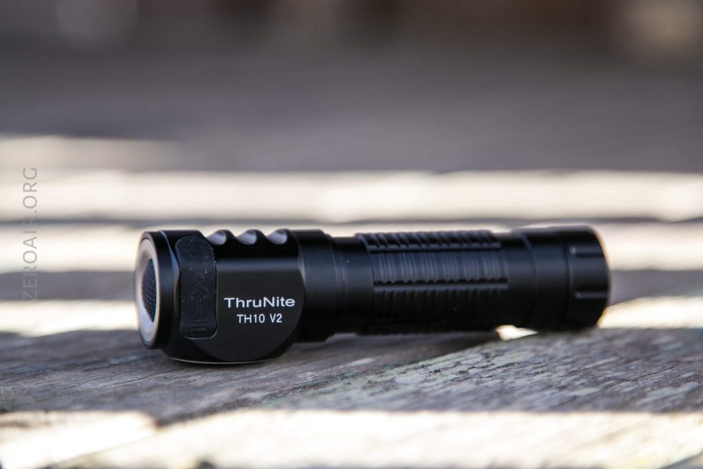 A black ThruNite TH10 V2 flashlight is lying on its side on a wooden surface, with clear branding and model name visible on the flashlight body.