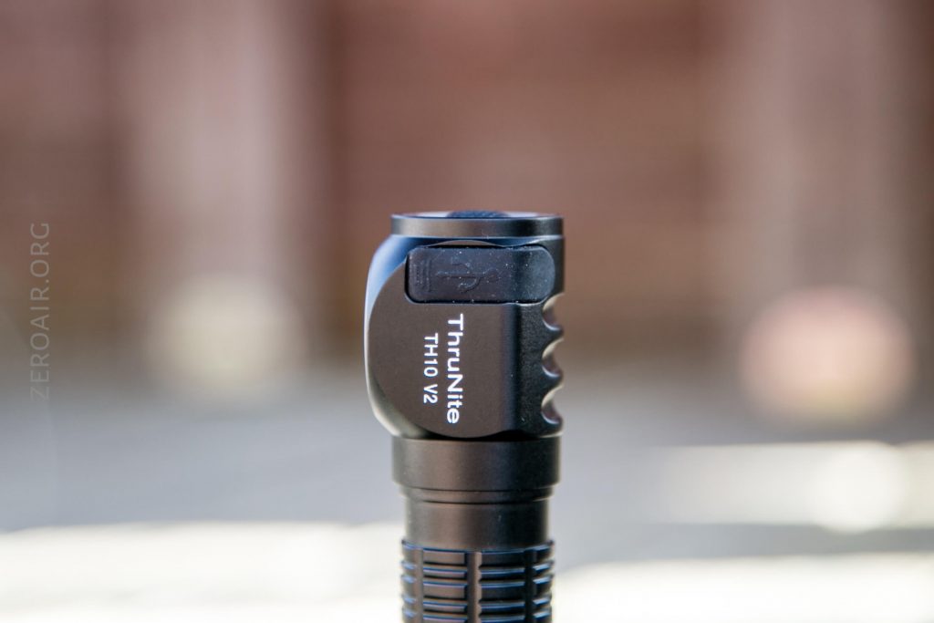 Close-up of a black ThruNite TH10 V2 flashlight standing upright outdoors with a blurred background. The product name and logo are clearly visible on the flashlight head.