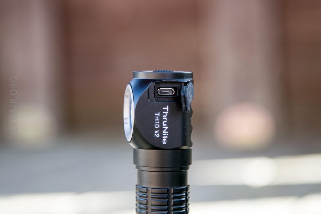 A close-up of a black ThruNite TH10 V2 flashlight standing upright, displaying its side button, USB charging port, and the model name printed on its body. The background is softly blurred.