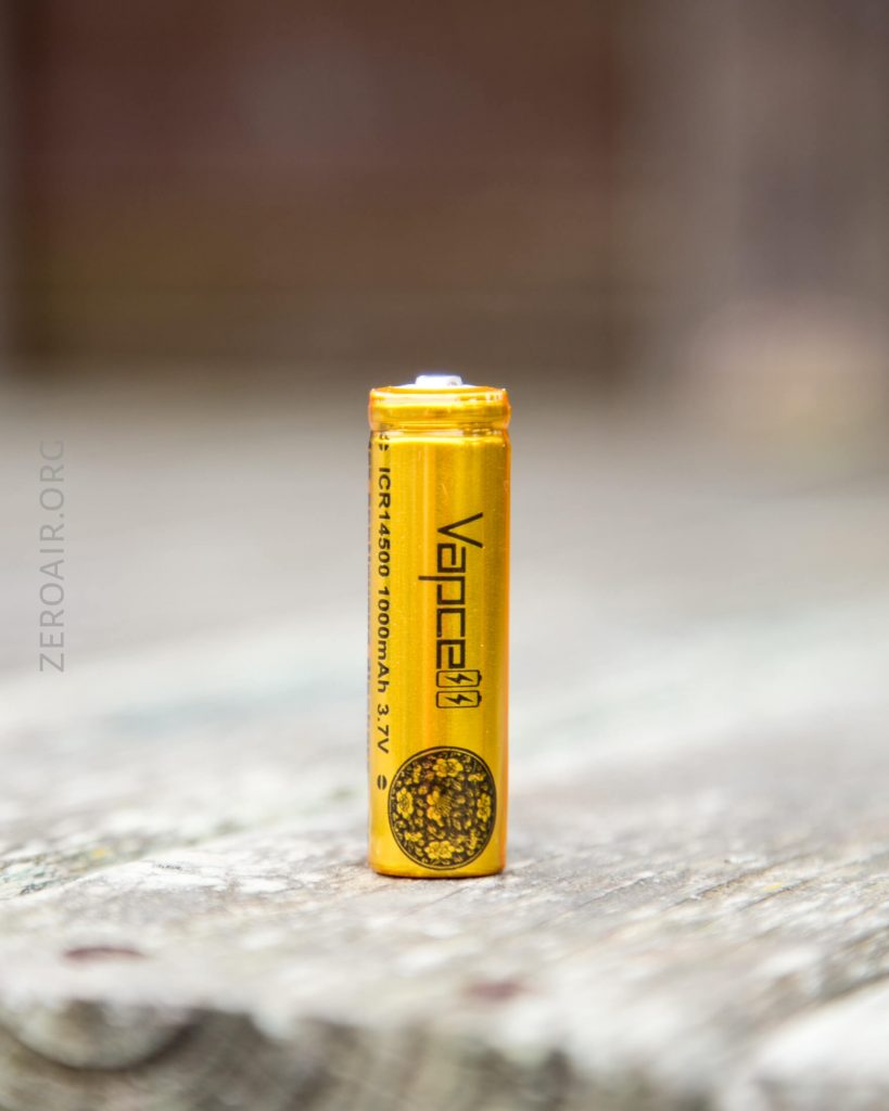 A gold VAPCELL 1000mAh 3.7V rechargeable battery stands upright on a weathered wooden surface, with a blurred background. The brand name and specifications are printed on the batterys side.