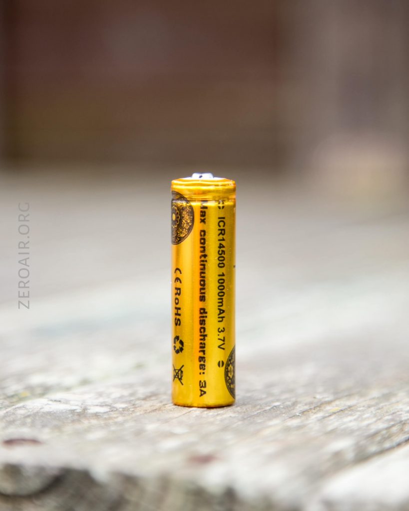 A yellow cylindrical battery is standing upright on a wooden surface. The battery label reads “ICR14500 1000mAh, 3.7V” along with other technical information and certification logos.