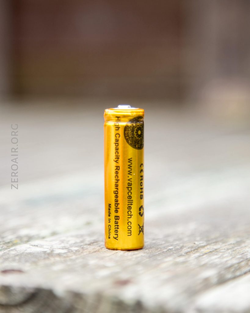 A single yellow AA rechargeable battery stands upright on a weathered wooden surface. The battery has black text and symbols printed on it. The background is blurred.