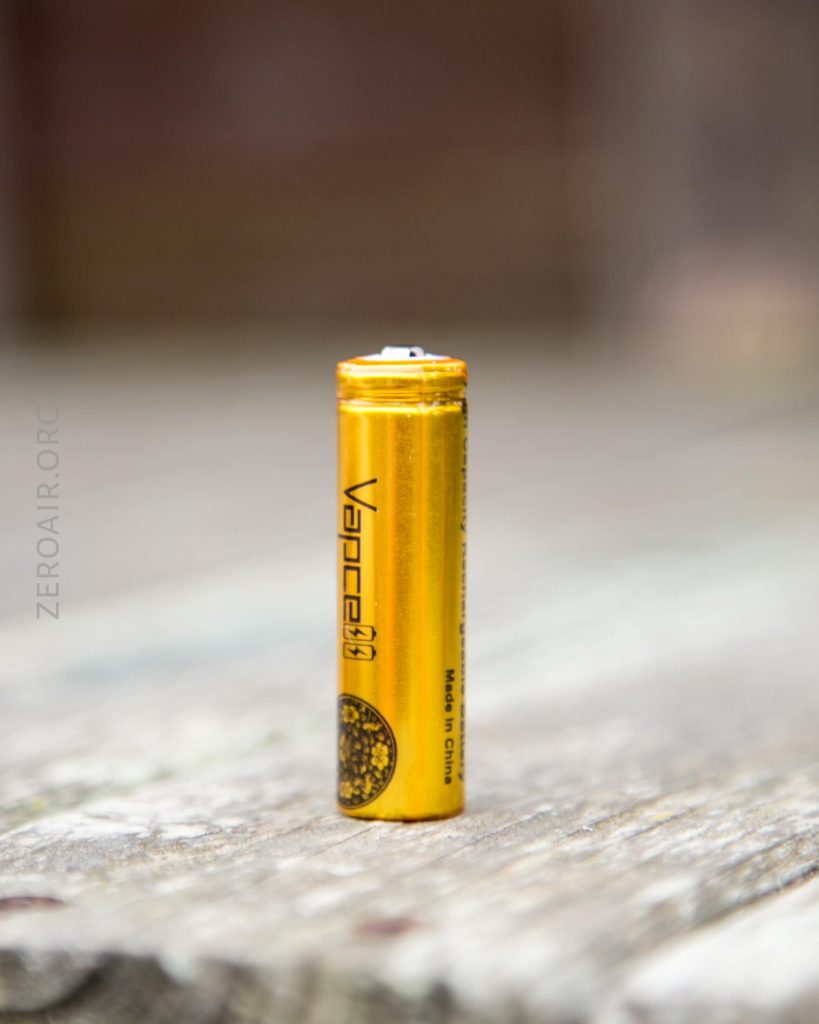A single yellow Vapcell AAA battery stands upright on a weathered wooden surface, with blurred background and faint ZEROAIR.ORG text along the left side.