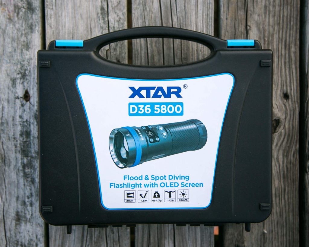 A black XTAR-branded carrying case with a label showing an image of the D36 5800 flashlight, described as a flood and spot diving flashlight with OLED screen, on a wooden surface.