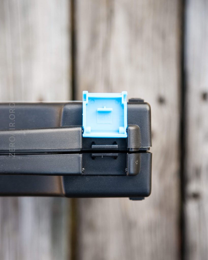 Close-up of a blue mechanical keyboard switch mounted on the corner of a black keyboard case, placed on a wooden surface.