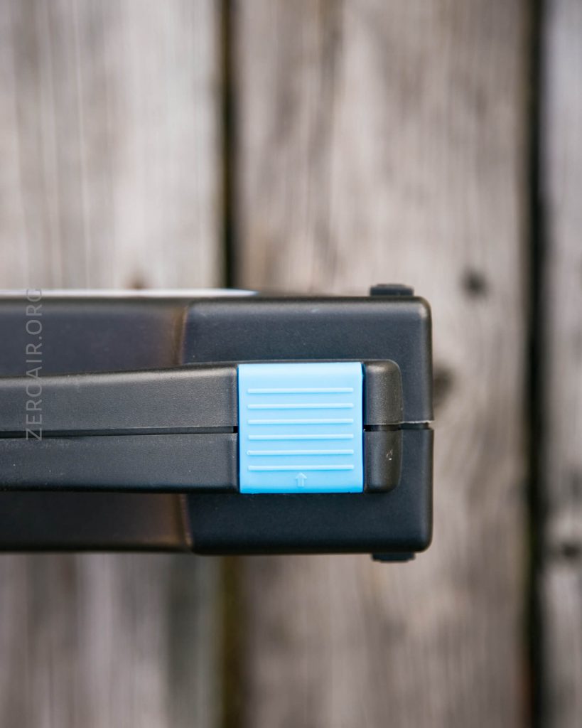 Close-up of the corner of a black plastic case with a blue, textured latch. The case is placed on a weathered wooden surface with visible grain and knots.