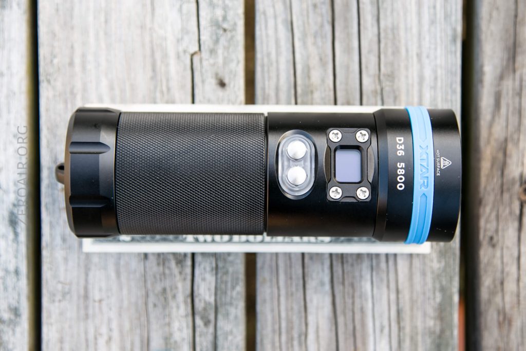 A black and blue XTAR D36 5800 flashlight is placed horizontally on top of a white rectangular object, resting on a weathered wooden surface. The flashlight features buttons and a small display screen.