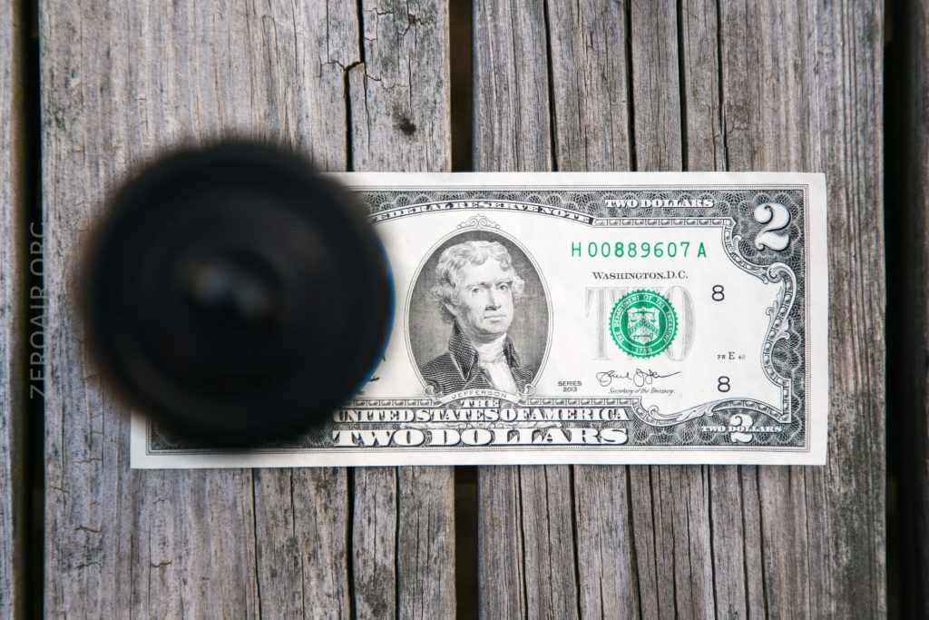 A two-dollar bill lies flat on a wooden surface, partially obscured by a black round object placed on the left side of the bill.