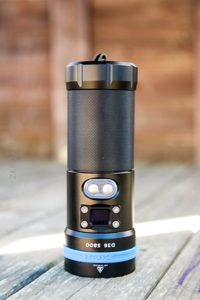 A black flashlight with a textured grip and blue accent stands upright on a wooden surface. The flashlight has two small lights and a digital display panel on its body. The background is blurred wood.