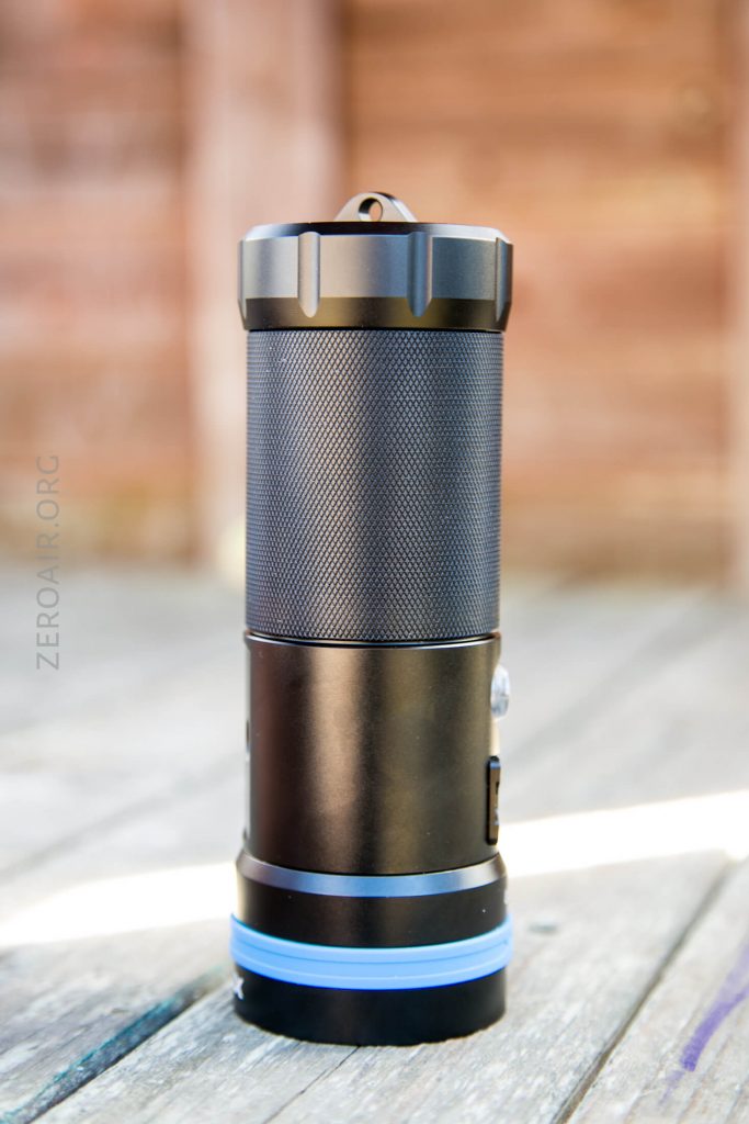 A black cylindrical flashlight with a textured grip and blue ring near the base, standing upright on a wooden surface with a blurred wooden background.