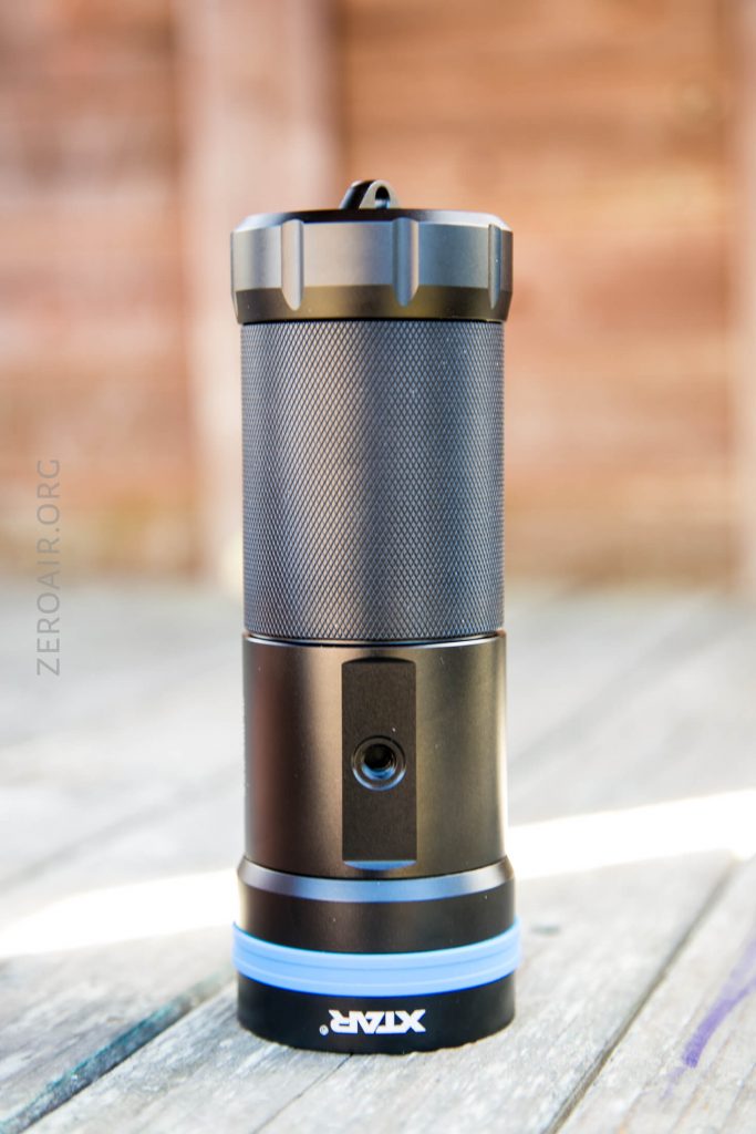 A black cylindrical flashlight with a textured grip and blue base stands upright on a wooden surface; the word “XTAR” is visible on the base. The background is out of focus.