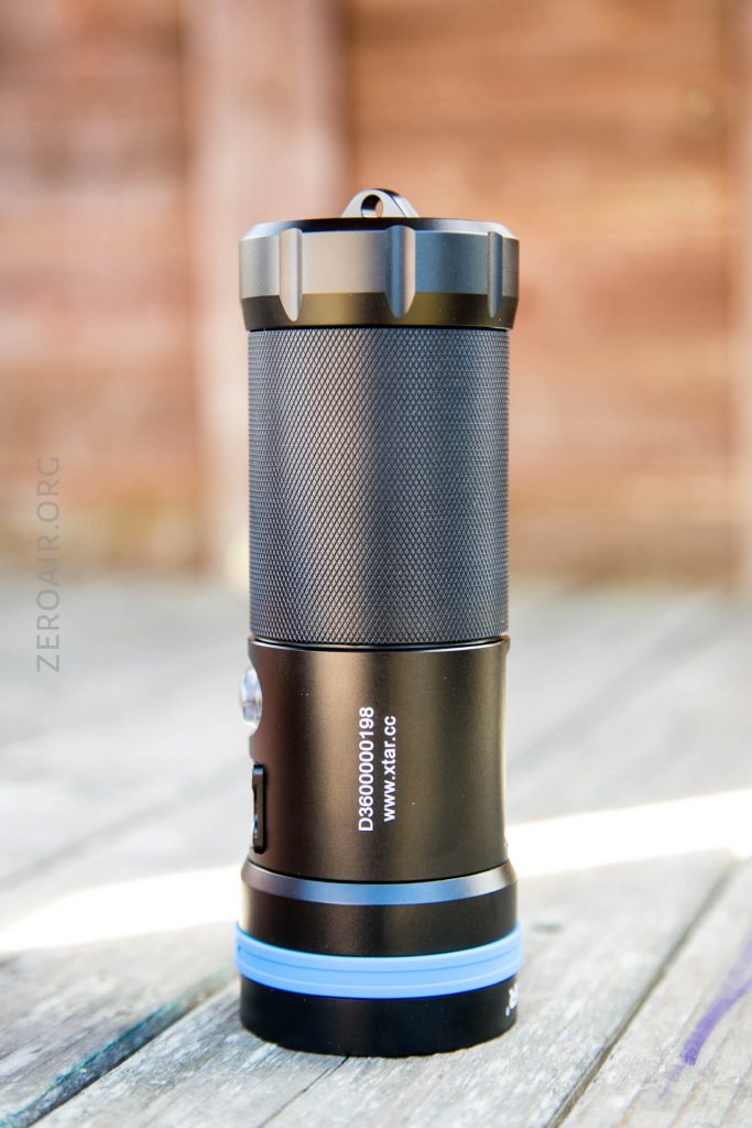 A black flashlight with a textured grip and blue ring at the base stands upright on a wooden surface. The flashlight has a button and a website address printed on its body. The background is blurred.