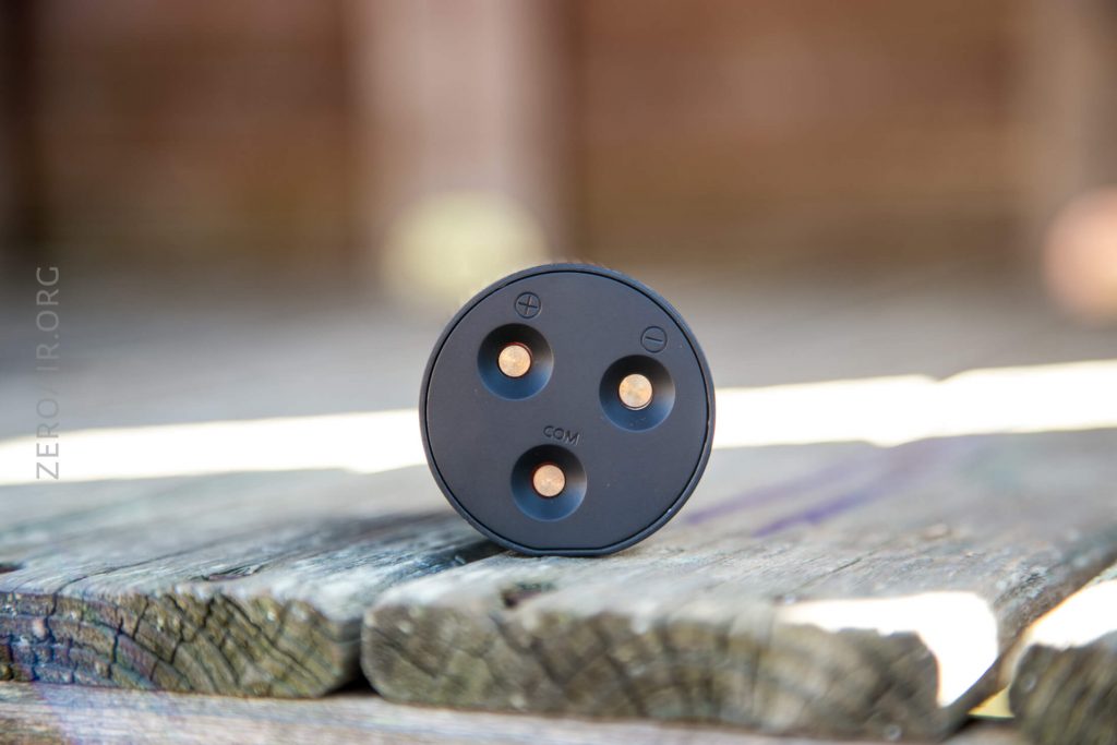 A round, black electrical connector with three circular metal prongs labeled with plus, minus, and COM symbols, placed upright on a weathered wooden surface.