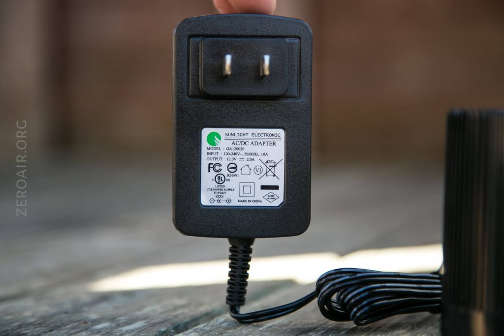 A hand holding a black Sunlight Electronic AC/DC adapter with a visible label showing input, output, certification marks, and voltage details. The adapter has two prongs and a coiled black cable.