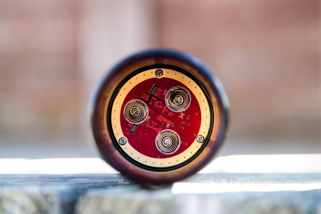Close-up view of the bottom of an electronic device showing a red circuit board with three visible metal coils and several small components, framed by a circular metallic edge.