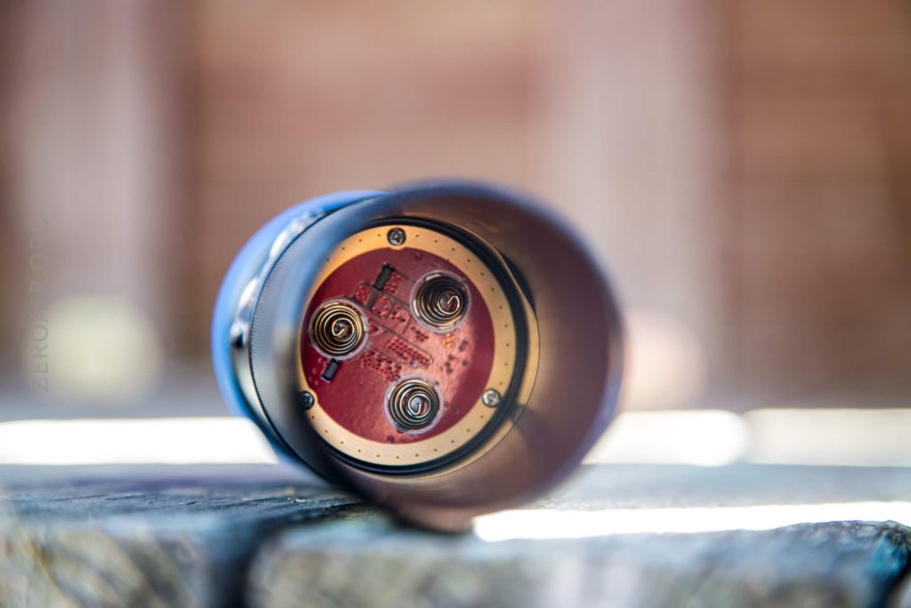 Close-up of the inside of a flashlight showing a red circuit board and three metal springs, with the flashlight lying on its side on a wooden surface. The background is out of focus.