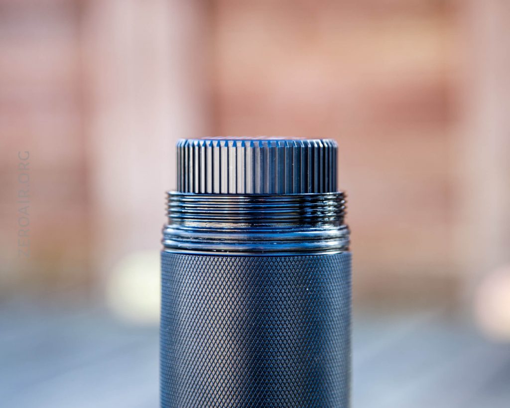 A close-up of a metallic cylindrical object with a textured grip and threaded top, shown partially unscrewed against a blurred background.