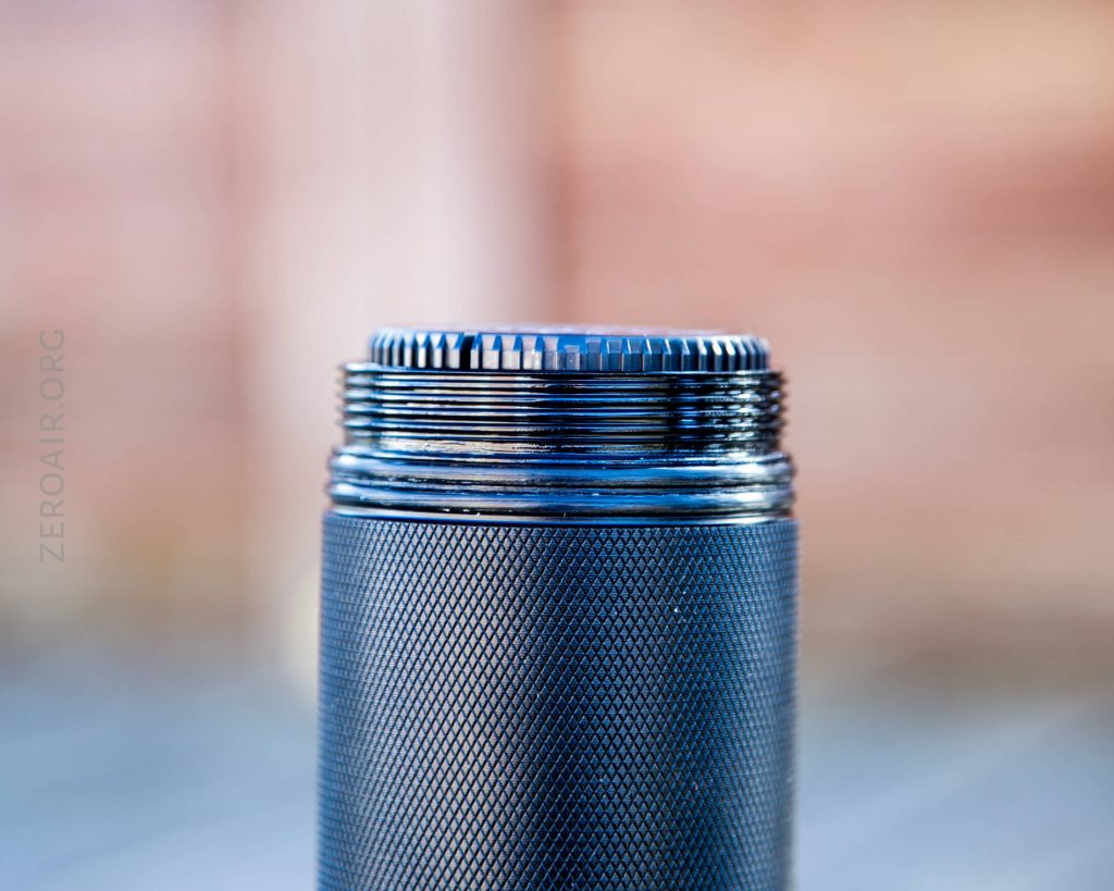 A close-up of a metal cylindrical object with a threaded top and textured grip, possibly a flashlight body, against a blurred background. The text ZEROAIR.ORG is faintly visible on the left side.