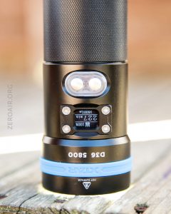 A close-up of a black and blue flashlight standing upright on a wooden surface, displaying its control panel and digital screen that reads “227 lm” and “2:21.” The brand “Xtar” and model “D36 5800” are visible.