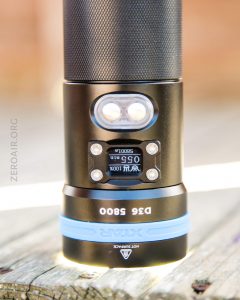 A close-up of a black and blue XTAR D36 5800 flashlight standing upright on a wooden surface, showing its digital display and buttons on the front. The background is blurred.