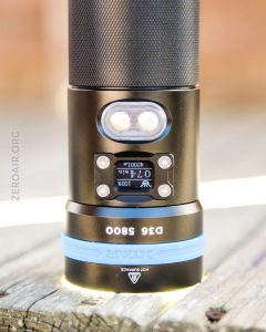 A close-up of a black and blue flashlight labeled Xtar D26 5800 with a digital display screen showing 074 and two round buttons above the display. The background is blurred.