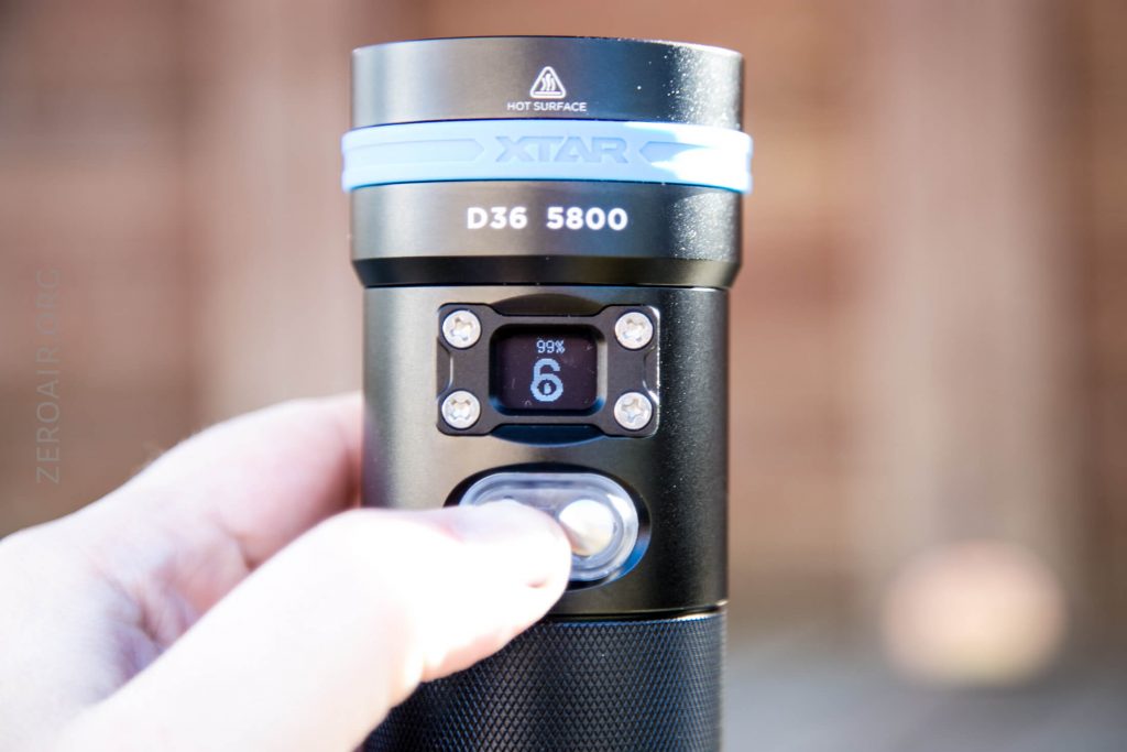 A hand holds a black XTAR D36 5800 flashlight, with a display screen showing “99%” and “6.” The flashlight has a blue ring near the top and visible control buttons below the screen.