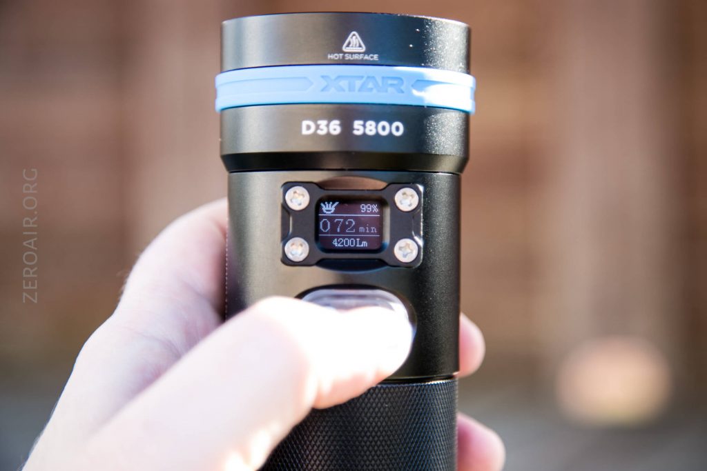 A close-up of a hand holding an XTAR D36 5800 flashlight, showing its digital display reading 072 min, 99%, and 4200 lm. The flashlight is black with a blue ring near its top.