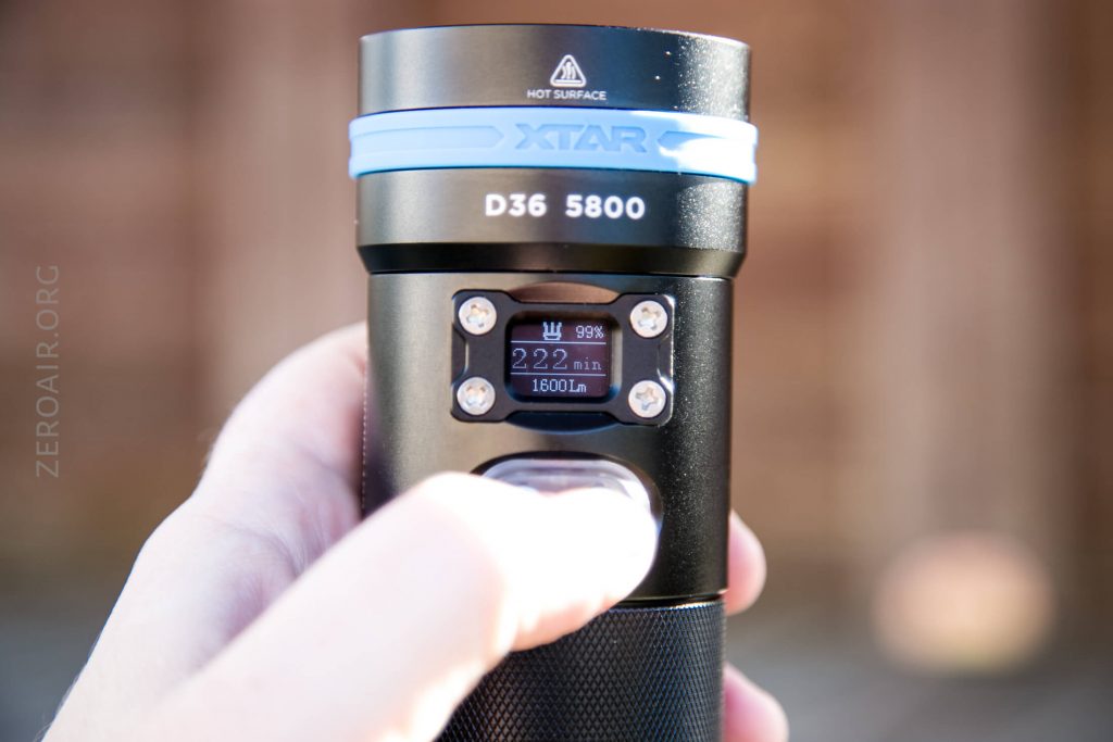 A hand holds an XTAR D36 5800 flashlight. Its display screen shows 222 min remaining, 1600 lm, and a battery icon at 99%. The flashlight also has a HOT SURFACE warning label near the top.