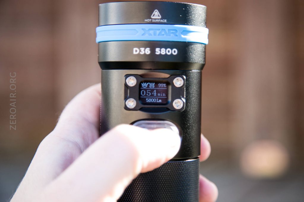 A hand holds a black XTAR D36 5800 flashlight, with a small display screen showing 054 min, 5800 Lm, and a 99% battery icon. The background is blurred.
