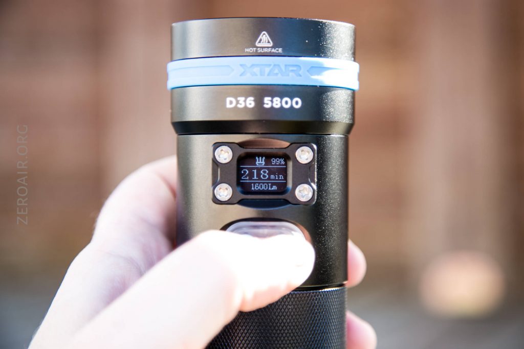 A hand holds an XTAR D36 5800 flashlight, displaying a screen that shows 99% battery, 218 minutes, and 1600 lumens. The words HOT SURFACE and branding are visible near the flashlight head.