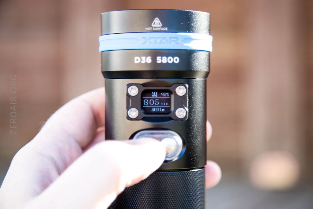A hand holding and turning on a black XTAR D36 5800 flashlight; the small display screen shows settings, including a runtime of 805 minutes at 400 lumens with a battery indicator at 99 percent.