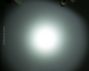 A bright white light beam shines in the center of a grayish wall with ZEROAIR.ORG faintly visible on the left side; two ceiling light fixtures appear partially at the top right corner.