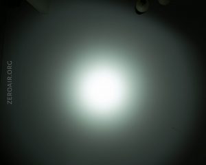 A bright white light shines on a dark wall, creating a circular glow with a fading edge; part of a ceiling light fixture is visible in the top right corner, and ZEROAIR.ORG is faintly written on the left side.