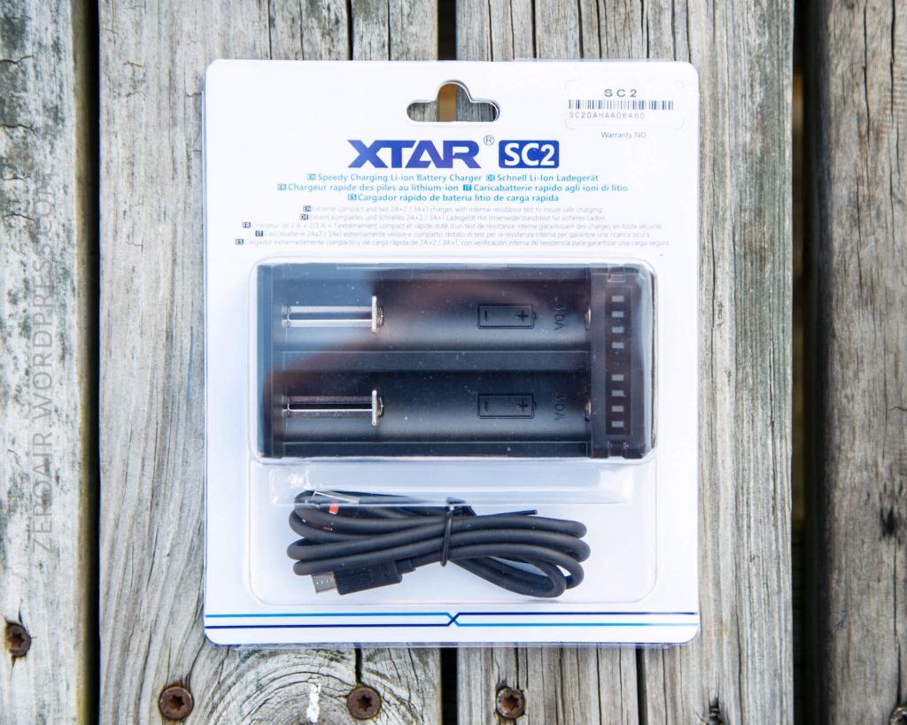 A packaged XTAR SC2 battery charger with two slots and an included USB cable, displayed on a weathered wooden surface. The packaging shows product information in multiple languages.