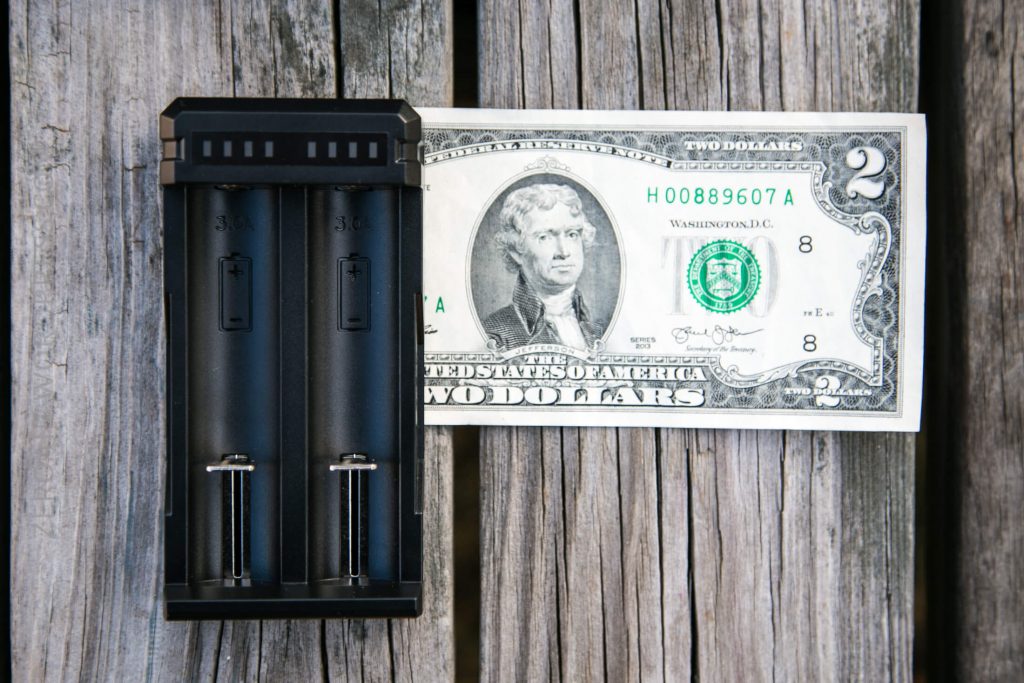 A black battery charger with two empty slots is placed on weathered wooden boards next to a two-dollar bill featuring Thomas Jefferson.
