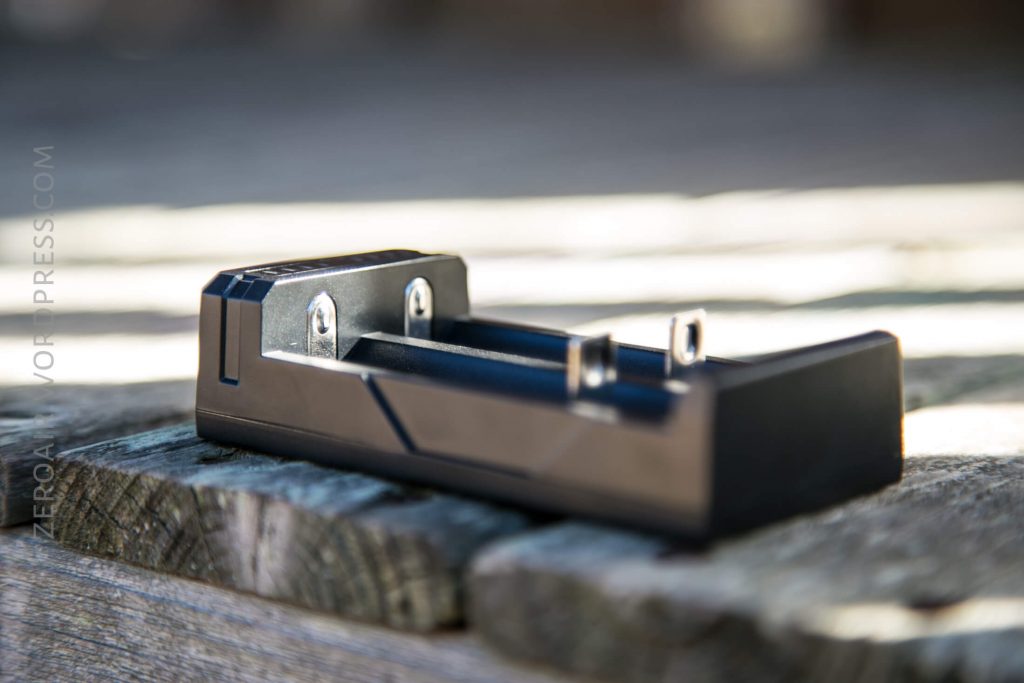 A black battery charger with metal charging contacts is placed on a weathered wooden surface outdoors, with sunlight casting shadows in the background.
